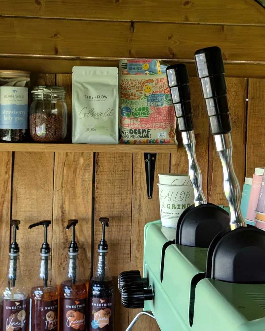 A bag of Fire & Flow roasted beans, syrups and branded cups on the shelf beside the Astoria machine